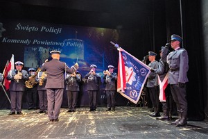 Zdjęcie na którym na scenie znajdują się policjanci pocztu sztandarowego będzińskiej komendy oraz policjanci Orkiestry Komendy Wojewódzkiej Policji w Katowicach.