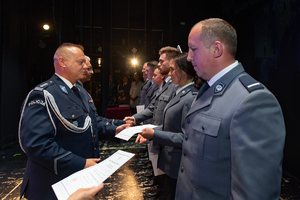 Komendant Powiatowy Policji w Będzinie w asyście Zastępcy Komendanta Wojewódzkiego grupie policjantów i policjantce na scenie akty mianowania.