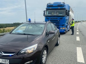 Na zdjęciu umundurowany policjant stoi przy samochodzie ciężarowym na poboczu drogi ekspresowej i rozmawia z kierowcą.