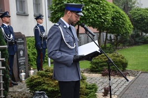 Zdjęcie przedstawia lektora prowadzącego uroczystość przy pomniku przed Komendą Powiatową Policji w Będzinie, za policjantem widoczni inni mundurowi tworzący wartę honorową.