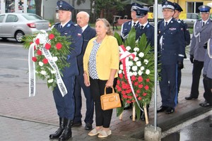 Na zdjęciu policjant trzyma wiązankę kwiatów, za nim stoi kobieta i mężczyzna a za nimi Komendant Wojewódzki Policji w Katowicach, Komendant Powiatowy Policji w Będzinie oraz inni mundurowi.