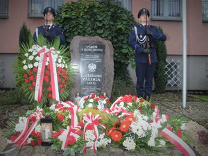 Na zdjęciu policjanci stoją obok pomnika tworząc wartę honorową. Przed pomnikiem znajdują się złożone kwiaty.