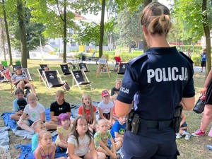 Na zdjęciu policjantka stoi przed grupą dzieci na dworze i przemawia do nich.