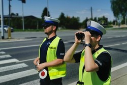 Na zdjęciu dwóch policjantów ruchu drogowego stoją przy drodze. Jeden trzyma w ręce tarczę do zatrzymywania pojazdów, a drugi lornetkę, przez którą patrzy.