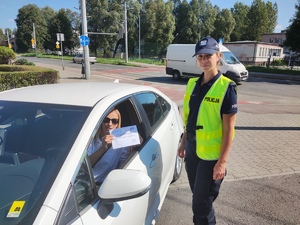 Zdjęcie. Policjantka stoi obok samochodu. W samochodzie znajduje się kobieta, która w ręce trzyma certyfikat wzorowego kierowcy.