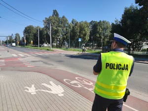 Na zdjęciu widzimy policjanta, który stoi  przy drodze z urządzeniem, które mierzy prędkość.