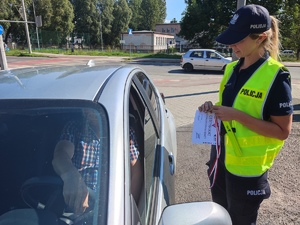 Zdjęcie, na którym policjantka stoi przy samochodzie i trzyma w rękach medal, certyfikat wzorowego kierowcy oraz odblask.