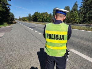 Na zdjęciu umundurowany policjant stoi z ręcznym miernikiem prędkości przy drodze.