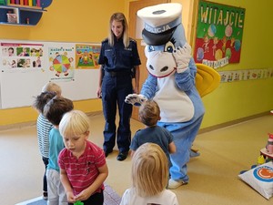 Na zdjęciu znajdują się dzieci które podchodzą do policjantki i maskotki policyjnej.