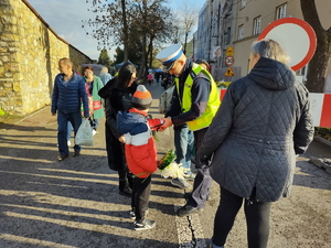 Zdjęcie. Umundurowany policjant zakłada opaskę odblaskową dziecku przy terenie cmentarza.