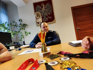 Zdjęcie przedstawiające puchar i medale zdobyte przez będzińskich policjantów, dalej widać siedzącego przy biurku w gabinecie Komendanta Powiatowego Policji w Będzinie inspektora Arkadiusza Więcka.