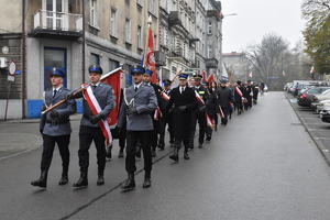 Zdjęcie, na którym widzimy przemarsz ludzi przez ulice miasta. Na czele idzie Sztandar Komendy Powiatowej Policji w Będzinie.