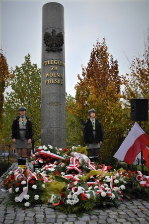 Zdjęcie, na którym widzimy dwie dziewczyny z harcerstwa ustawione przy pomniku &quot;Poległym za Wolną Polskę&quot; poniżej znajdują się wiązanki kwiatów.