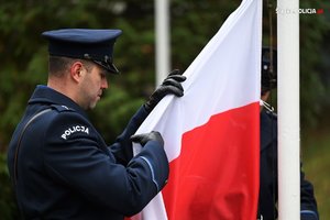 Na zdjęciu policjant trzyma w rękach flagę, która wciągana jest na maszt.