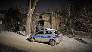 Zdjęcie, na którym stoją policjanci przy radiowozie i opuszczonym budynku.