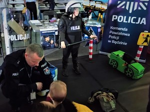 Zdjęcie, na którym widzimy policjanta, który pomaga małemu dziecku założyć na siebie imitację samochodu. Dalej widzimy dziecko przebrane za policjanta.