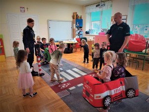 Na zdjęciu policjantka i policjant uczą grupę dzieci przepisów ruchu drogowego wykorzystując mobilne miasteczko ruchu drogowego w sali.