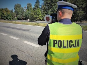 Na zdjęciu policjant drogówki stoi przy drodze i miedzy prędkość za pomocą urządzenia pomiarowego.