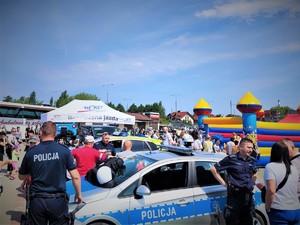 Na zdjęciu policjanci rozmawiają z ludźmi podczas motoryzacyjnej imprezy, na której znajduje się wielu fanów motoryzacji.