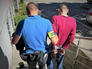 Na zdjęciu policjant prowadzi zatrzymanego, który ma założone kajdanki na ręce trzymane z tyłu.