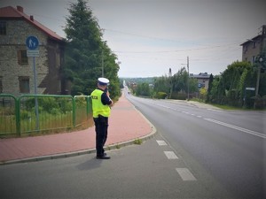 Na zdjęciu policjant stoi przy drodze i z urządzeniem pomiarowym mierzy prędkość.