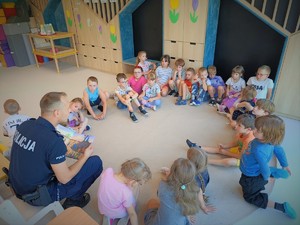 Na zdjęciu policjant siedzi w kole z dziećmi i czyta im książkę.