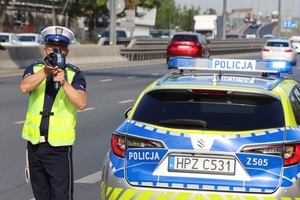 Na zdjęciu policjant stoi przy radiowozie i mierzy prędkość urządzeniem pomiarowym.