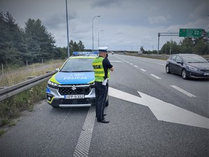 Na zdjęciu policjant przy radiowozie mierzy prędkość urządzeniem pomiarowym.