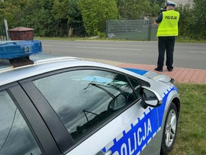 Na zdjęciu radiowóz policyjny oraz policjant stojący przy drodze z urządzeniem pomiarowym do mierzenia prędkości.