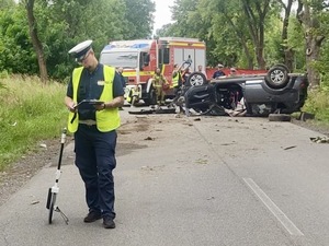 zdjęcie kolorowe przedstawiające umundurowanego policjanta podczas wykonywania oględzin, a za nim rozbite pojazdy na skutek wypadku