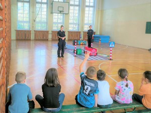 Na zdjęciu policjantki stoją przed grupą dzieci na sali gimnastycznej i odpowiadają na pytania.