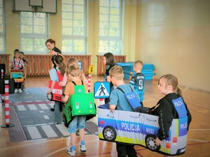 Na zdjęciu dzieci są poprzebierane za różne pojazdy i ćwiczą naukę przepisów przy policjantkach.