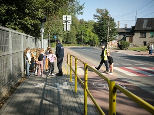 Na zdjęciu policjant i policjantka z grupą dzieci przy przejściu dla pieszych.