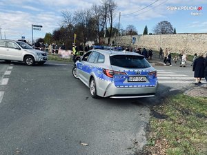 Zdjęcie. Policjant przed radiowozem kieruje ruchem w okolicy cmentarza, gdzie przemieszczają się ludzie.