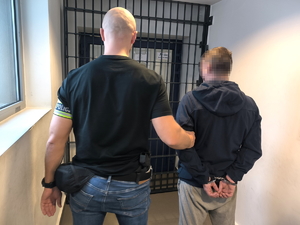 Na zdjęciu policjant pionu kryminalnego stoi przed kratą z zatrzymanym, który ma założone kajdanki na ręce trzymane z tyłu.