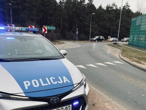 Na zdjęciu radiowóz, z tyłu widoczny policjant, który bada kierujących na drodze.