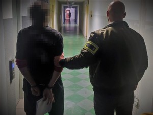 Na zdjęciu nieumundurowany policjant z opaską z napisem POLICJA trzyma zatrzymanego na korytarzu, który ma założone kajdanki na ręce trzymane z tyłu.