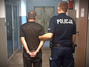 Na zdjęciu policjant trzyma zatrzymanego, który ma założone kajdanki na ręce trzymane z tyłu.