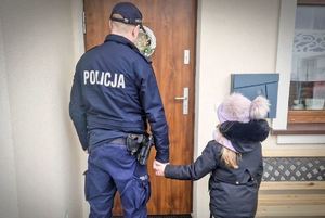 Zdjęcie, na którym mundurowany policjant trzyma za rękę dziewczynkę. Obydwoje stoją przed drzwiami.