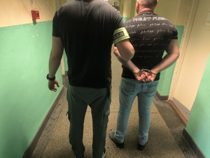 Na zdjęciu policjant trzyma zatrzymanego mężczyznę, który ma założone kajdanki na ręce trzymane z tyłu.