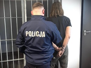 Na zdjęciu policjant z zatrzymanym mężczyzną, który ma założone kajdanki na ręce trzymane z tyłu.
