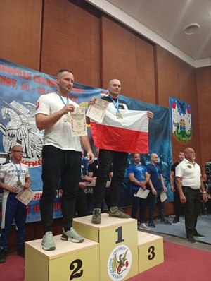 Na zdjęciu stoją mężczyźni na podium. Zwycięzca trzyma biało-czerwoną flagę.