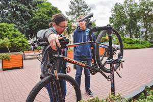 Na zdjęciu kobieta przy rowerze, który znajduje się na stojaku. Za nią stoi chłopiec.