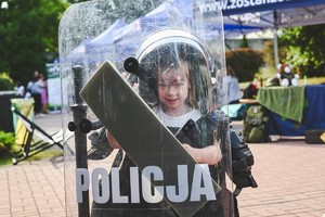 Na zdjęciu mała dziewczynka w białym kasku trzyma tarczę policyjną.