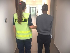Na zdjęciu policjantka trzyma zatrzymanego, który ma kajdanki założone na ręce trzymane z przodu w policyjnym areszcie.