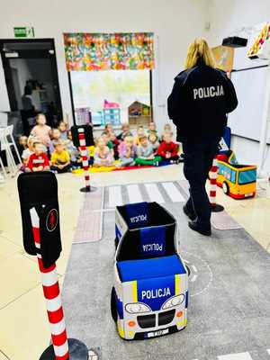 Na zdjęciu grupa policjantka prowadzi prelekcje z przepisów ruchu drogowego dla grupy przedszkolaków.