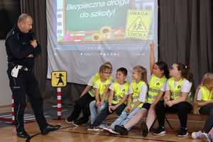 Na zdjęciu policjant z mikrofonem stoi przy grupie uczniów, którzy mają kamizelki odblaskowe.