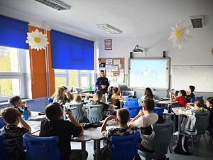 Na zdjęciu policjantka stoi w klasie i mówi do uczniów, którzy siedzą w ławkach.