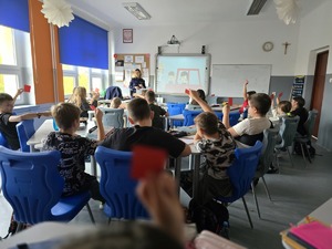 Na zdjęciu policjantka stoi w klasie i mówi do uczniów, którzy trzymają w rękach kartki koloru czerwonego i zielonego.