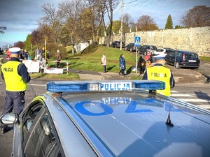 Na zdjęciu dwóch policjantów ruchu drogowego stoją przy radiowozie w okolicy cmentarza.